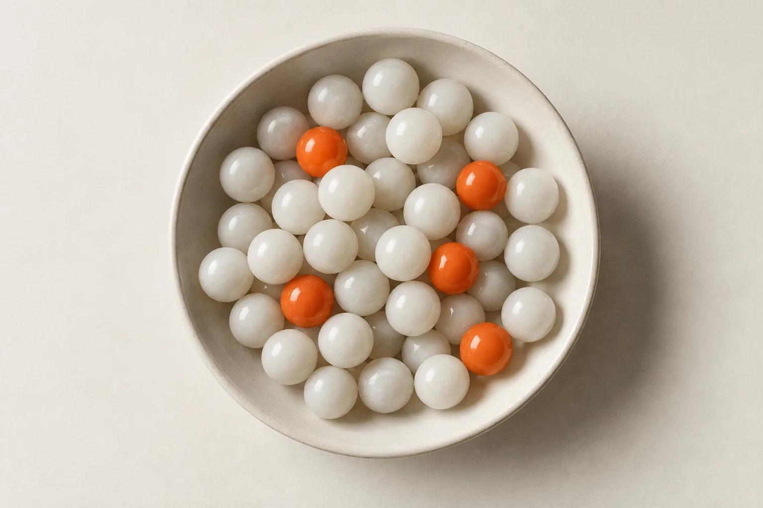 A white bowl of white marbles with a few orange ones among them.